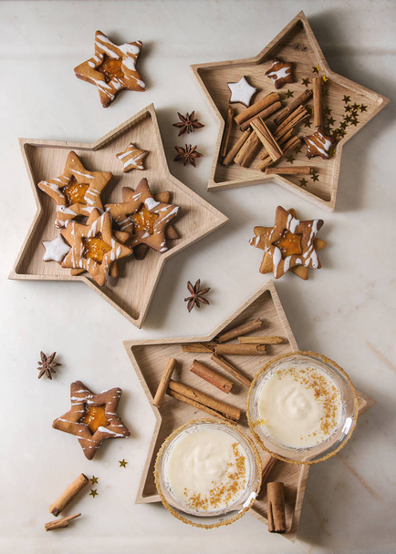Eggnog Christmas milk cocktail, served in two vintage crystal glasses on wooden tray with shortbread star shape sugar cookies, cinnamon sticks over white marble background. Flat lay, space - 写真・画像