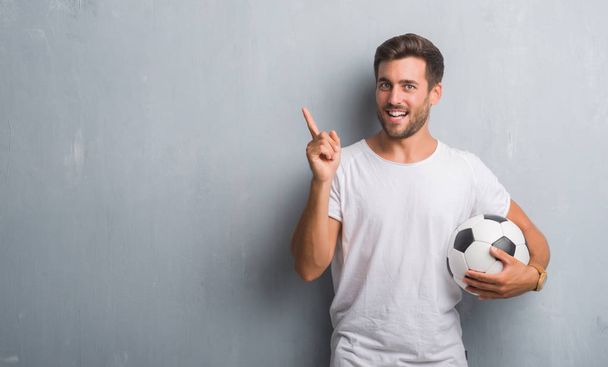 Csinos, fiatal férfi, gazdaság foci futball labda nagyon boldog, mutat, a kéz és az ujjak oldalán szürke grunge falon - Fotó, kép