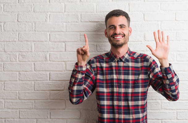 Fiatal felnőtt férfi fehér tégla falon mutatja, és ujjal felfelé mutató állandó száma hat, miközben mosolyogva, magabiztos és boldog. - Fotó, kép