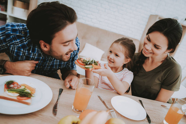 Girl Gives Father Sandwich. Family Breakfast. Family at Table. Fruit on Plate. Sandwich on Plate. White plate. Juice in a glass. Food on a table. Happy Family. Smiling People. Vegetables on Board. - Fotó, kép