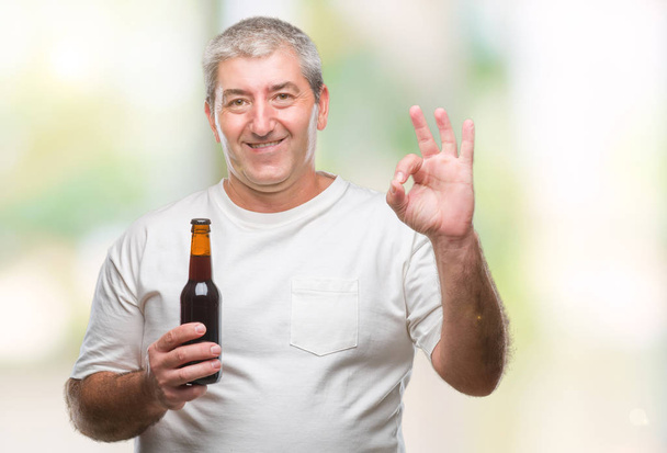 Bonito homem sênior bebendo garrafa de cerveja sobre fundo isolado fazendo sinal ok com os dedos, excelente símbolo
 - Foto, Imagem