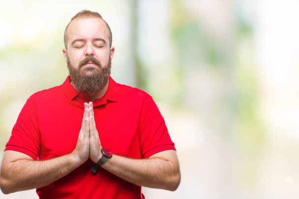 Молодой кавказский хипстер в красной рубашке на изолированном фоне молится руками, прося прощения, уверенно улыбаясь.
. - Фото, изображение