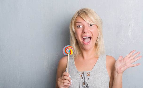 Kaukasische volwassen vrouw over grijs grunge muur eten snoep lollipop erg blij en opgewonden, winnaar expressie vieren overwinning schreeuwen met grote glimlach en getogen handen - Foto, afbeelding
