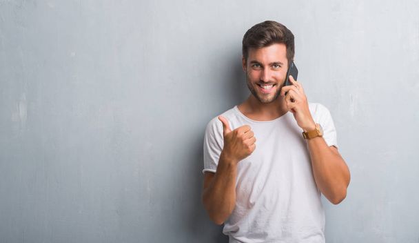 Handsome young man over grey grunge wall speaking on the phone happy with big smile doing ok sign, thumb up with fingers, excellent sign - Foto, Bild
