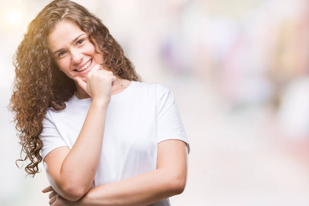 Beautiful brunette curly hair young girl wearing casual t-shirt over isolated background looking confident at the camera with smile with crossed arms and hand raised on chin. Thinking positive. - Foto, afbeelding