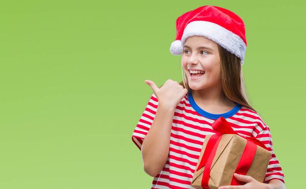 Young beautiful girl wearing christmas hat and holding gift over isolated background pointing and showing with thumb up to the side with happy face smiling - Photo, Image
