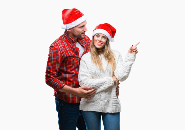 Молода пара закохана в різдвяний капелюх над ізольованим тлом з великою посмішкою на обличчі, вказуючи рукою і пальцем в сторону, дивлячись на камеру
. - Фото, зображення