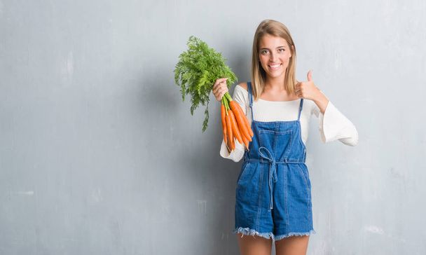 Beautiful young woman over grunge grey wall holding fresh carrots happy with big smile doing ok sign, thumb up with fingers, excellent sign - Photo, Image