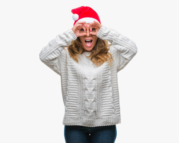Mujer hispana mayor de mediana edad con sombrero de navidad sobre un fondo aislado haciendo un gesto aceptable como binoculares sacando la lengua, ojos mirando a través de los dedos. Expresión loca
. - Foto, Imagen