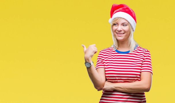 Jovem bela mulher loira vestindo chapéu de Natal sobre fundo isolado sorrindo com o rosto feliz olhando e apontando para o lado com o polegar para cima
. - Foto, Imagem