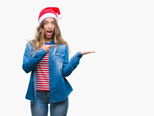 Красивая молодая блондинка в рождественской шляпе на изолированном фоне удивлена и улыбается в камеру, представляя с рукой и указывая пальцем
. - Фото, изображение