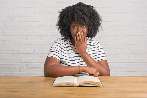 junge afrikanisch-amerikanische Frau sitzt auf dem Tisch und liest einen Buchumschlag Mund mit der Hand schockiert vor Scham für Fehler, Ausdruck von Angst, Angst in der Stille, geheimes Konzept - Foto, Bild
