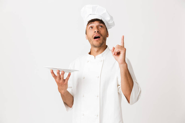 Image of shocked handsome young chef man standing isolated over white wall background pointing to copyspace holding plate. - Fotografie, Obrázek