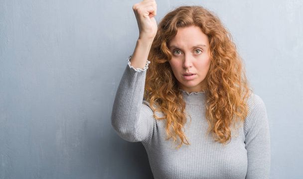 Молодая рыжая женщина через серую гранж стену раздражен и разочарован кричать от гнева, сумасшедший и кричать с поднятой рукой, гнев концепции
 - Фото, изображение
