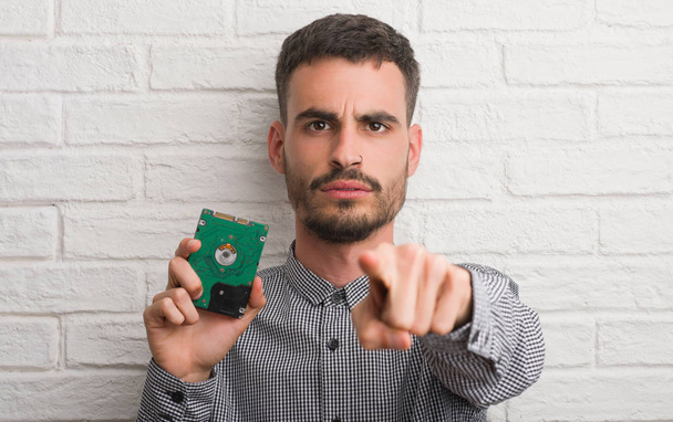 Young adult man over brick wall holding hard drive pointing with finger to the camera and to you, hand sign, positive and confident gesture from the front - Photo, image