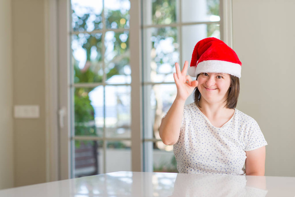 Σύνδρομο Down γυναίκα στο σπίτι φορώντας καπέλο Χριστουγέννων κάνει εντάξει πινακίδα με τα δάχτυλα, εξαιρετική σύμβολο - Φωτογραφία, εικόνα