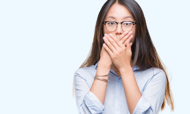 Молодая азиатская деловая женщина в очках на изолированном фоне шокировала закрывая рот руками за ошибку. Секретная концепция
. - Фото, изображение