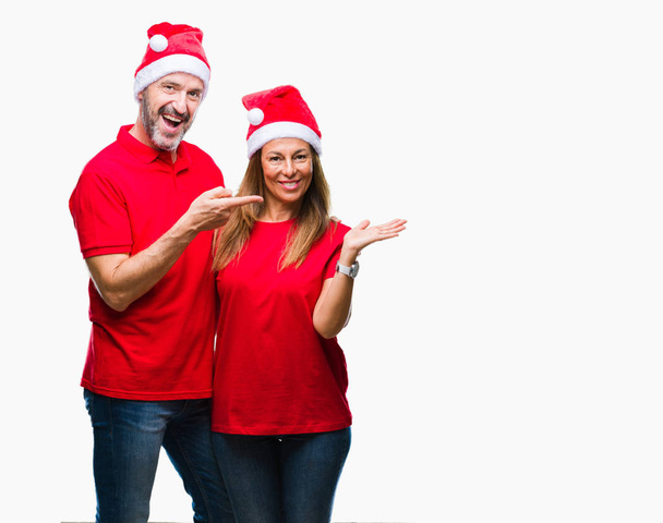 Hispanisches Paar mittleren Alters mit Weihnachtsmütze über isoliertem Hintergrund erstaunt und lächelnd in die Kamera, während es mit der Hand präsentiert und mit dem Finger zeigt. - Foto, Bild