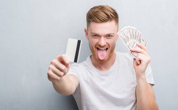 Rude młody człowiek gospodarstwa dolarów i karty kredytowej, trzymanie języka out z wyrażenie zabawny zadowolony. Emocja koncepcja. - Zdjęcie, obraz