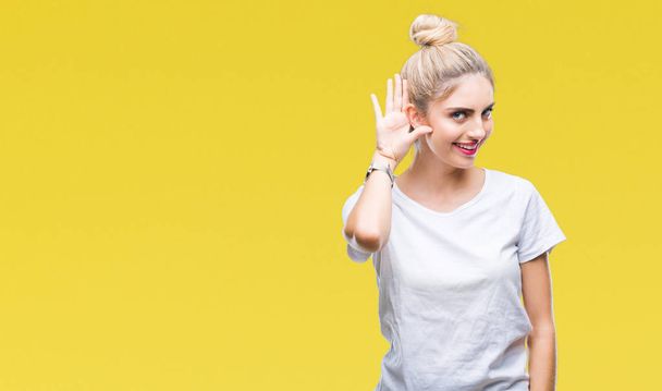 Mladá krásná blondýna žena nosí bílé tričko izolované pozadí s úsměvem s rukou přes ucho naslouchá slyšení zvěsti nebo drby. Hluchota koncept. - Fotografie, Obrázek