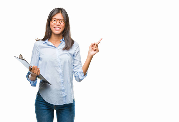 Jeune femme d'affaires asiatique sur fond isolé tenant presse-papiers très heureux pointant avec la main et le doigt sur le côté
 - Photo, image