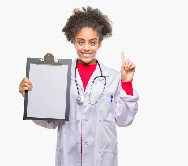 Jonge afro Amerikaanse dokter vrouw bedrijf klembord over geïsoleerd achtergrond verrast met een idee of vraag vinger met blij gezicht, nummer één - Foto, afbeelding
