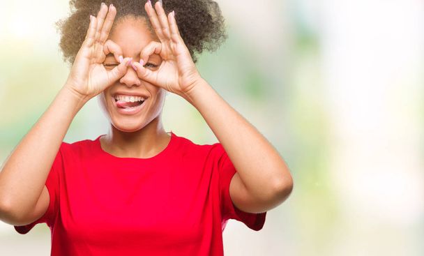 Young afro american woman over isolated background doing ok gesture like binoculars sticking tongue out, eyes looking through fingers. Crazy expression. - Photo, Image