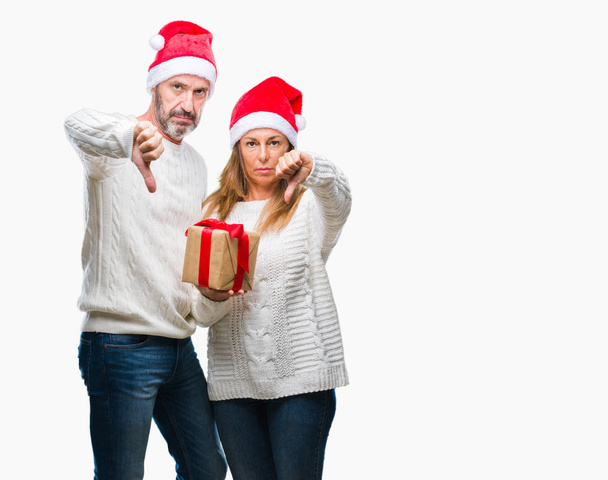 Hispánský pár středního věku klobouk vánoční a drží dar nad izolované pozadí s naštvaný obličej, záporné znaménko ukazující nechuť s palci dolů, koncept odmítnutí - Fotografie, Obrázek