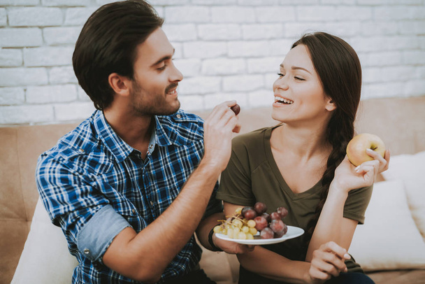 Happy Man and Woman. Man with Grapes on Plate. Man Feeding Grapes Woman. Family in Room. Smiling Family at Home. Male and Famale in Sofa. White Pillows in Brown Sofa. Smiling Person. White Plate. - Fotografie, Obrázek