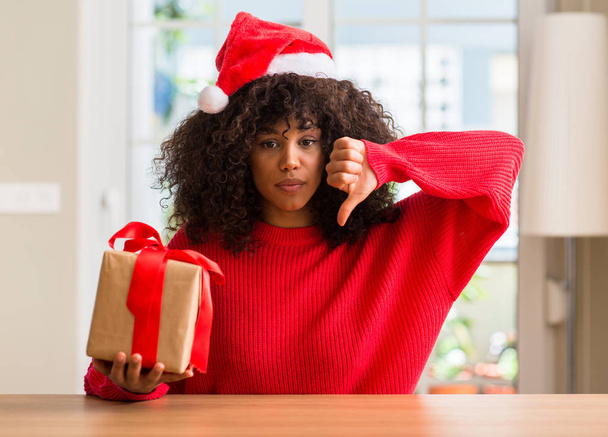 Mujer afroamericana sosteniendo presente vistiendo sombrero rojo navidad con cara enojada, signo negativo que muestra aversión con los pulgares hacia abajo, concepto de rechazo
 - Foto, imagen