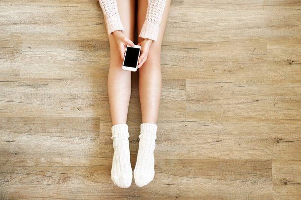 Lazy afternoon concept. Young woman wearing knee high socks, knitted sweater, sitting on wooden laminated floor, holding blank screen mobile phone. Background, copy space, top view, close up. - 写真・画像