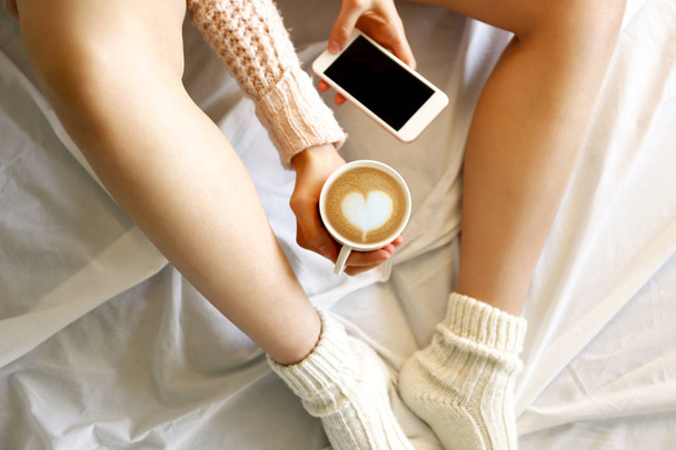 Lazy afternoon concept. Close up shot of woman sitting in bed, reading, holding red cup of cappuccino coffee with heart shaped latte art foam & blank screen mobile phone. Background, copy space. - Foto, imagen