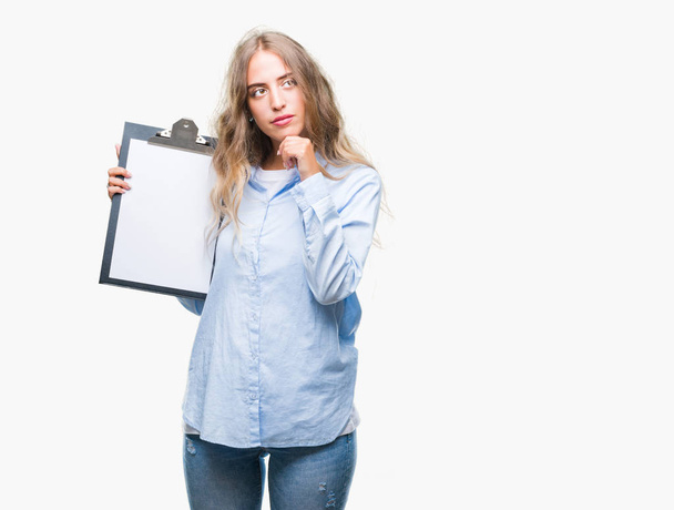 Beautiful young blonde business woman holding clipboard over isolated background serious face thinking about question, very confused idea - Фото, изображение