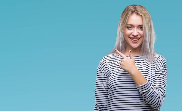 Young blonde woman over isolated background cheerful with a smile of face pointing with hand and finger up to the side with happy and natural expression on face looking at the camera. - Foto, imagen