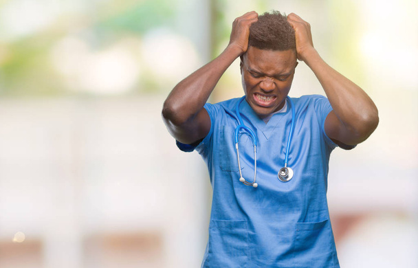 Młody lekarz african american mężczyzna na białym tle sobie chirurg jednolite cierpiących na bóle głowy, zdesperowany i podkreślił, bo bóle i migreny. Ręce na głowę. - Zdjęcie, obraz