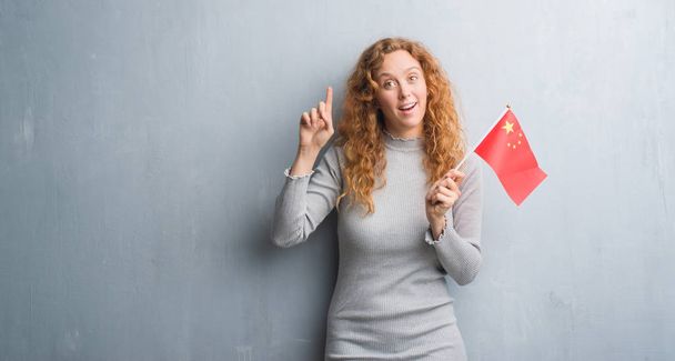 Young redhead woman over grey grunge wall holding flag of China surprised with an idea or question pointing finger with happy face, number one - Φωτογραφία, εικόνα