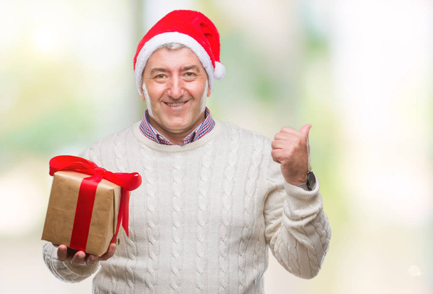 Komea vanhempi mies yllään joulu hattu ja pitämällä lahja yli eristetty tausta osoittaa ja osoittaa peukalolla sivulle iloinen kasvot hymyillen
 - Valokuva, kuva