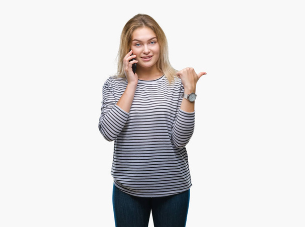 Mujer joven caucásica mostrando la pantalla del teléfono inteligente sobre un fondo aislado apuntando y mostrando con el pulgar hacia el lado con la cara feliz sonriendo
 - Foto, imagen