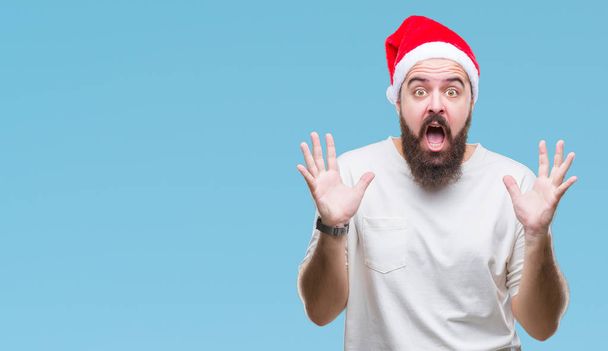上クリスマスの帽子を着ている若い白人ヒップ男クレイジーを祝う背景を分離した腕を上げると成功のためびっくりし、叫んで興奮して目を開きます。勝者の概念 - 写真・画像