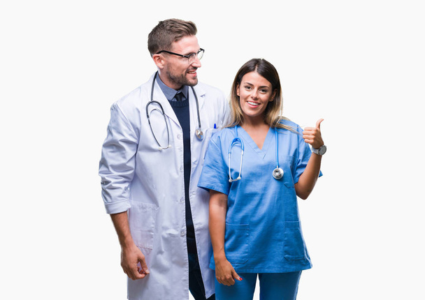 Νεαρό ζευγάρι γιατρός και χειρούργος πέρα από το απομονωμένο υπόβαθρο χαμογελώντας με χαρούμενο πρόσωπο αναζητούν και τοποθετώντας το δείκτη στην πλευρά με τον αντίχειρα. - Φωτογραφία, εικόνα