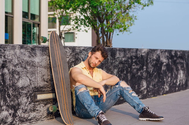 Young handsome man with beard sitting with longboard on the street. - Foto, immagini