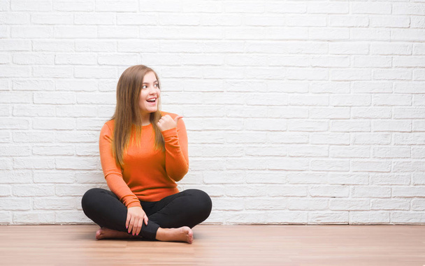 Fiatal felnőtt nő ül a földön az őszi fehér tégla falon mutató és hüvelykujjával, akár a boldog arc mosolyogva oldalon mutatja - Fotó, kép