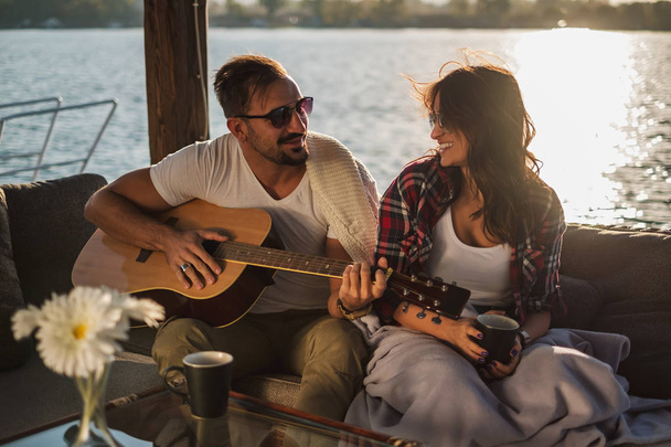 Homem tocando guitarra para sua namorada junto ao rio enquanto ela está sorrindo. Belo pôr do sol
 - Foto, Imagem