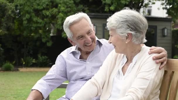 Senior couple sitting together on bench at park. Elderly married couple sitting outdoor and relaxing. Romantic husband embrace his wife while looking away and smiling. Future and retirement concept. - Footage, Video