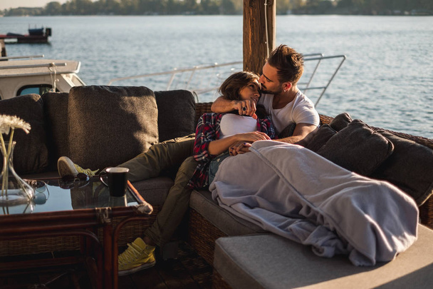 Novio besando a su novia en la frente mientras ella está acostada cubierta con manta junto al río. Hermoso día de otoño
 - Foto, Imagen
