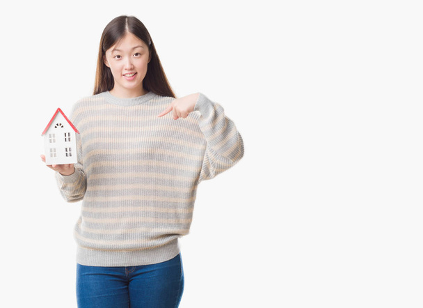 Молодая китайская реальная государственная агент женщина на изолированном фоне держит дом с неожиданным лицом указывая пальцем на себя
 - Фото, изображение