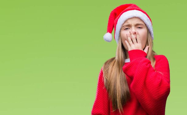 Молодая красивая девушка в рождественской шляпе на изолированном фоне скучает зевая усталый закрывающий рот рукой. Беспокойство и сонливость
. - Фото, изображение