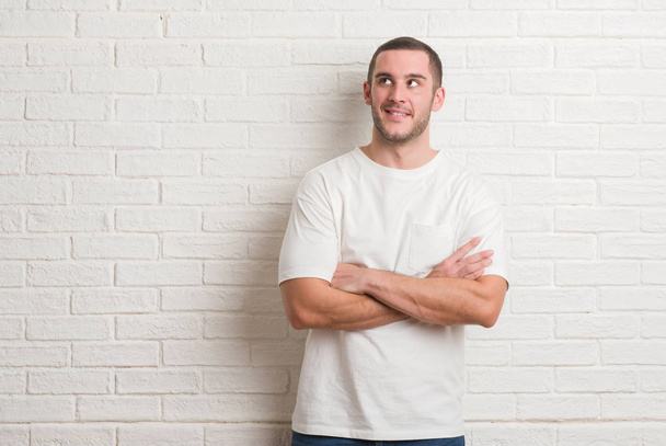 Jeune homme caucasien debout sur un mur de briques blanches souriant côté regardant et regardant loin en pensant
. - Photo, image
