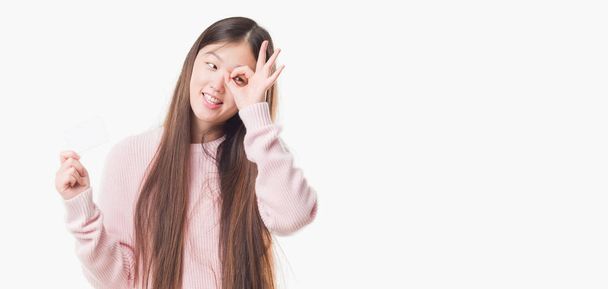 Jeune femme chinoise sur fond isolé tenant carte de visite avec le visage heureux souriant faire ok signe avec la main sur les yeux regardant à travers les doigts
 - Photo, image
