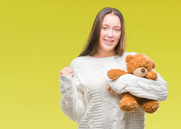 Young caucasian woman holding teddy bear over isolated background screaming proud and celebrating victory and success very excited, cheering emotion - Фото, изображение